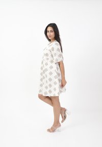 Abito bianco con motivo geometrico beige, maniche corte e design fluido. Indossato con sandali trasparenti con tacco per un look estivo leggero.