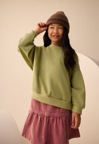 Grøn sweatshirt med lange ærmer og ribbet kant, kombineret med en lyserød kordnær nederdel med en vid bund. Brun strikket hue på hovedet.