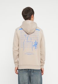 Beige hoodie with a blue graphic design featuring a table and plants. It has a drawstring hood and ribbed cuffs with a relaxed fit.