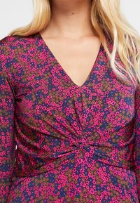 Kwiatowa sukienka z głębokim dekoltem w kształcie litery V, długimi rękawami i zebranym węzłem w okolicy talii. Materiał łączy kolory różowy, niebieski i zielony.