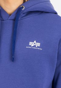 Lila huvtröja i bomull med dragsko i huooden. Den har en vit Alpha Industries-logotyp som är tydligt placerad på bröstet.