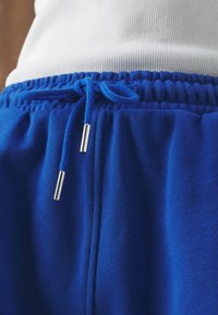 Modré tepláky mají texturovanou látku, elastický pas a nastavitelnou šňůrku s kovovými koncovkami. Detailní pohled zdůrazňuje prošívání.