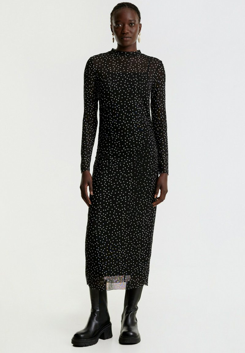 Schwarzes Langarmkleid mit weißen und bunten Punkten, figurbetonte Silhouette, durchsichtiges Material, knielang, getragen mit schwarzen Stiefeletten.