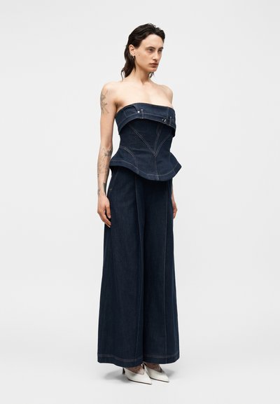 Nő sötétkék pánt nélküli farmer fűző felsőben és bő szárú nadrágban, fehér hegyes sarkú cipővel, világos egyszínű háttér előtt áll.