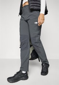 Osoba ubrana w ciemnoszare spodnie turystyczne The North Face, czarne buty turystyczne, białe skarpetki i czarną kamizelkę, stojąca na jednolitym tle.