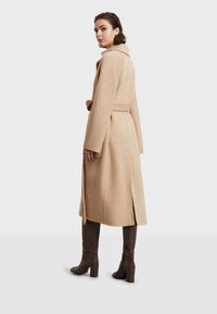 Abrigo de mezcla de lana beige con cinturón en la cintura, amplio cuello con muescas y aberturas laterales, combinado con botas altas marrones de tacón.