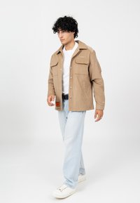 Jasnobrązowa kurtka z zapięciem na guziki, dwoma kieszeniami na piersi i teksturowanym wykończeniem. W połączeniu z jasnoniebieskimi dżinsami i białymi sneakersami.