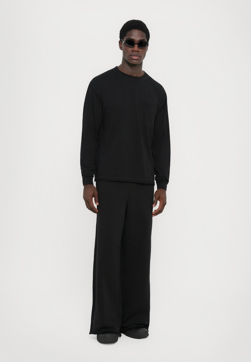 Chemise noire à manches longues avec une poche, assortie à un pantalon noir ample. Des chaussures plates et des lunettes de soleil complètent la tenue monochrome.