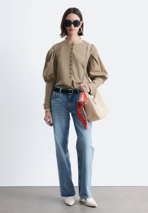 Kobieta w beżowej bluzce z bufkami, niebieskich dżinsach, białych butach na szpic, czarnych okularach przeciwsłonecznych, złotej biżuterii, nosząca beżową torebkę na ramię z czerwonym szalikiem.