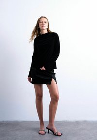 Une femme se tient contre un mur blanc, portant un haut à manches longues en velours noir, une mini-jupe noire fendue, et des sandales à talons noires ornées de strass.