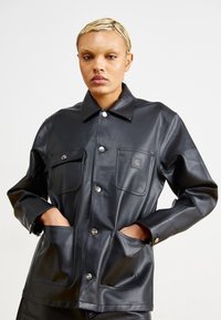 Giubbotto in pelle nera con design strutturato, colletto e due tasche frontali. Presenta bottoni argentati e una patch con logo sulla tasca sul petto.