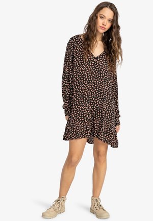 Joven mujer con cabello largo y ondulado, vistiendo un vestido negro de manga larga con estampado floral y botas beige hasta el tobillo con cordones, posando relajadamente.