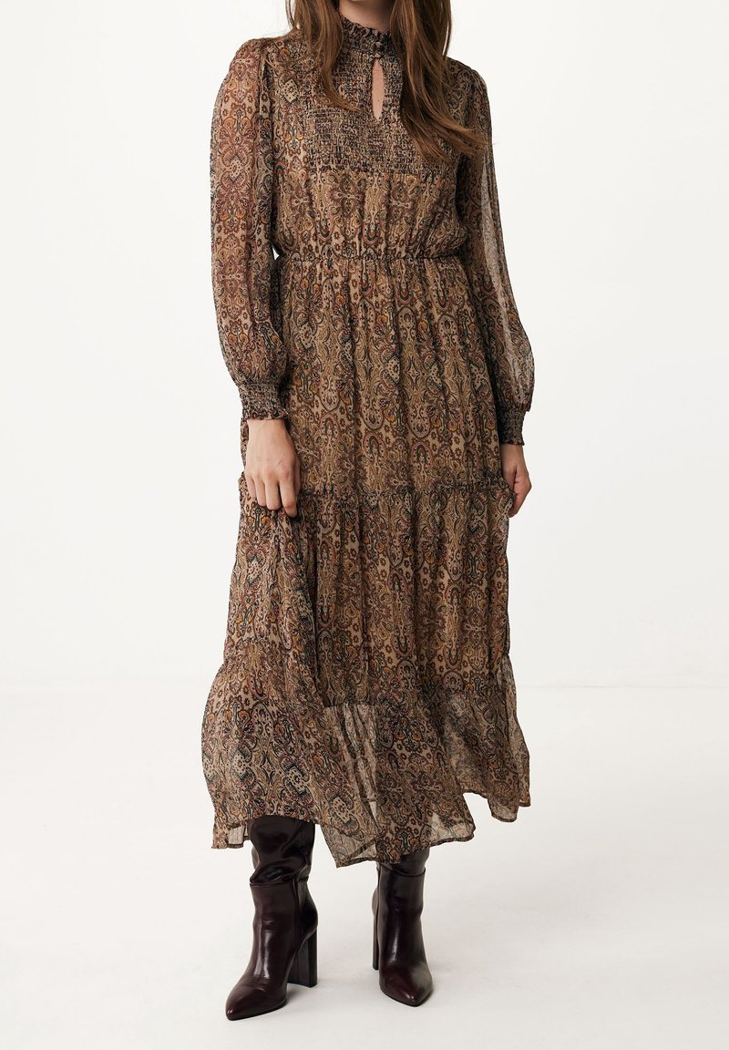 Femme portant une longue robe marron à motifs cachemire avec un col montant et des poignets froncés, associée à des bottes à talons marron foncé.