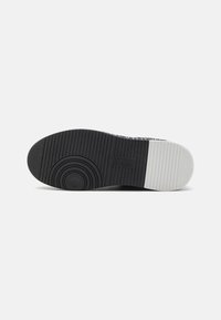 Semelle de chaussure en caoutchouc noir et blanc avec des motifs de bande de roulement circulaires et linéaires, montrant le texte de la marque embossé près de la zone de la voûte plantaire.