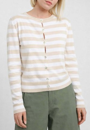 Donna che indossa un cardigan a righe beige e bianche con bottoni e maniche lunghe, e pantaloni verde oliva, su uno sfondo chiaro e uniforme.