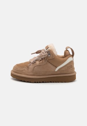 Tansuede en textielsneaker met een gestructureerde bovenkant, gevoerde kraag en rubberen zool. Heeft veters en een witte accent. UGG-logo zichtbaar.