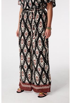 Pantalon à jambes larges avec motifs géométriques noirs, crème et rouille, taille nouée, accompagné de sandales noires ornées d'accents circulaires dorés.