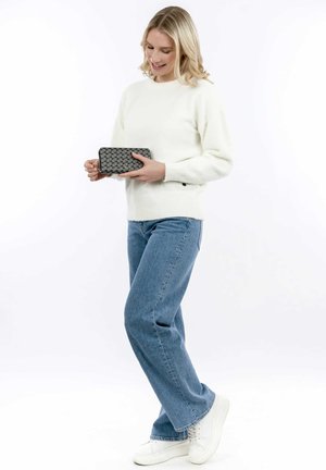 Biały puszysty sweter, niebieskie dżinsy i białe sneakersy. Trzyma ciemnoszary portfel o teksturowanym wzorze. Pozycja stojąca, jasne tło.