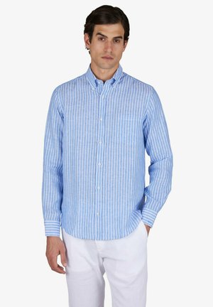 Homme portant une chemise à rayures verticales bleu clair et blanc à boutons, manches longues, et un pantalon blanc, debout avec une main dans la poche.