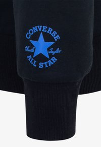 Cappotto nero con polsini che presentano un logo blu con una stella e il testo "CONVERSE ALL STAR Chuck Taylor". Texture a costine sui polsini.