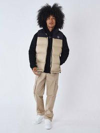 Gilet imbottito beige con collo nero, indossato sopra un felpa nera. I pantaloni cargo crema e le sneakers bianche completano l'outfit. Tessuto testurizzato e cerniere visibili.