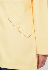 Κοντινό πλάνο σε τσέπη μπεζ-κίτρινης ζακέτας με καπάκι που κλείνει με κουμπί, δείχνοντας μέρος του μανικιού και ένα χέρι δίπλα της.