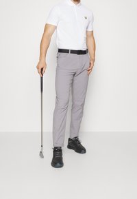 Mies, joka on pukeutunut valkoiseen poolopaitaan, harmaisiin housuihin, mustaan vyöhön ja mustiin kenkiin, pitää golfmailaa kädessään ja seisoo yksinkertaista valkoista taustaa vasten.