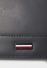 Portefeuille en cuir marine foncé présentant un logo rectangulaire avec des bandes rouges, blanches et noires. Texture lisse avec des détails de couture visibles.