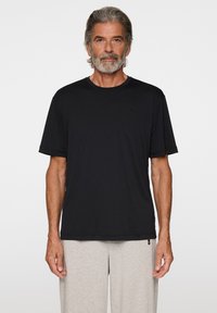 Man med grått hår och skägg klädd i en enkel svart t-shirt med kort ärm och ljusgråa mjukisbyxor mot en vit bakgrund.