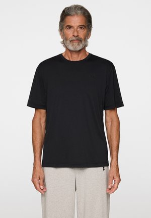 Homme aux cheveux et à la barbe grises portant un t-shirt noir à manches courtes uni et un pantalon de survêtement gris clair sur fond blanc.