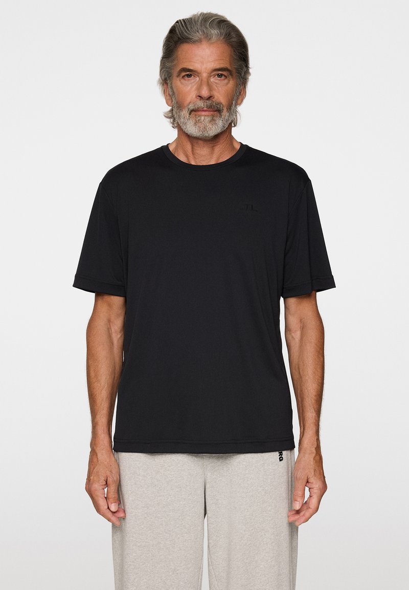 Man med grått hår och skägg klädd i en enkel svart t-shirt med kort ärm och ljusgråa mjukisbyxor mot en vit bakgrund.