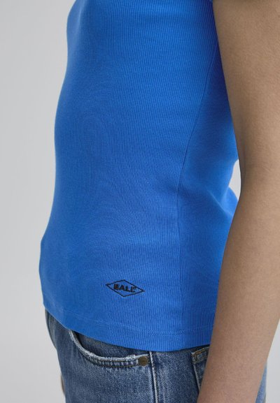 Mėlynas rievėtas maišelis su prigludusia kirpimo linija ir juodu logotipu apačioje; derinamas su suplėšytais džinsiniais šortais.