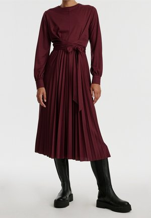 Robe plissée bordeaux à manches longues et ceinture à nouer, en tissu lisse ; associée à des bottes noires à plateforme montantes jusqu'aux genoux.