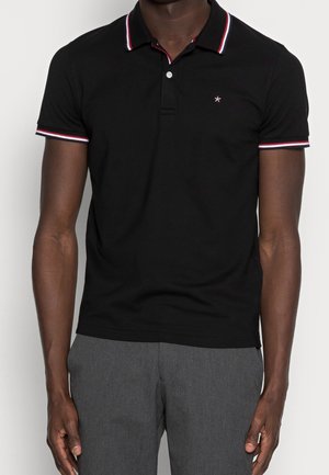 Homme portant un polo noir ajusté avec un col et des manches rayés rouges et blancs, assorti à un pantalon gris.