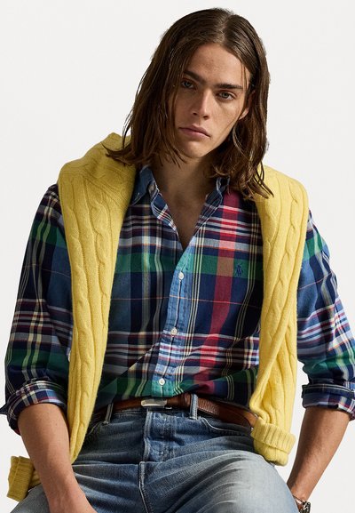 Geltonas megztas liemelis, dėvimas ant margaspalvio languoto marškinėlio, mėlyni džinsai ir ruda diržas. Tekstūriniai audiniai ir drąsūs raštai.