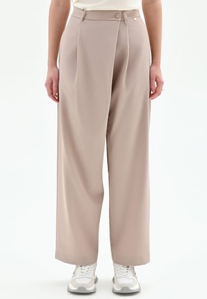 Pantaloni beige a vita alta con taglio a gamba larga, caratterizzati da pieghe frontali, chiusura con un solo bottone e una texture liscia. Indossati con sneaker bianche.