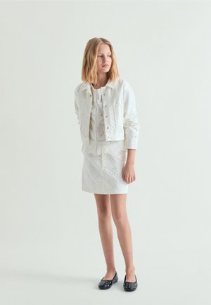 Dziewczyna o blond włosach do ramion, ubrana w białą kurtkę z ażurowym wzorem, pasującą spódnicę oraz czarne baleriny z ćwiekami, stojąca na jednolitym tle.