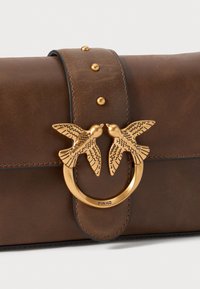 Bruine leren handtas met een ronde gouden hardware met twee vogelmotieven, geaccentueerd met decoratieve studs op de schouderband.