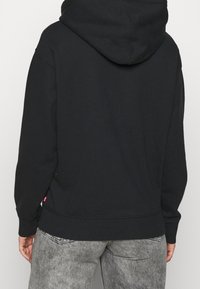 Persona che indossa una semplice felpa nera e jeans grigi sbiaditi, mostrata di spalle su uno sfondo neutro.