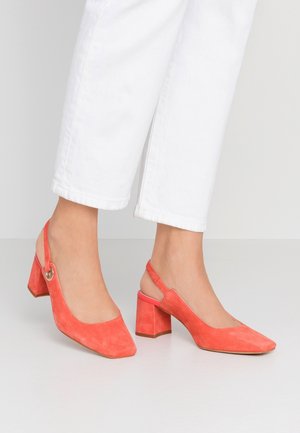 Stopy w koralowych zamszowych sandałach na słupku typu slingback, zestawione z białymi, krótkimi spodniami na neutralnym tle.