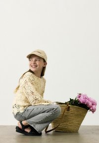 Maglione all'uncinetto giallo chiaro, jeans grigi, sandali neri e un cappello beige con la scritta "Good Vibes". Un cesto intrecciato con peonie rosa.