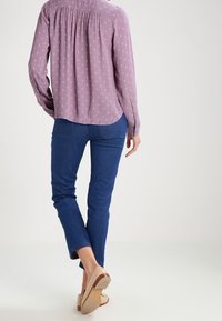 Person walking away wearing a long-sleeve purple patterned blouse, blue jeans, and beige shoes with low heels on a white background.