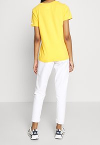 Camiseta de algodón amarilla con mangas cortas, combinada con pantalones blancos. El atuendo se completa con zapatillas deportivas con detalles en azul.