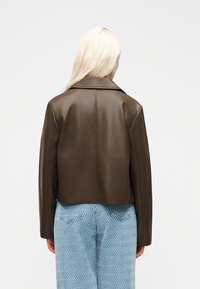 Person with long blonde hair wears a dark brown leather jacket and light blue patterned pants, standing facing away against a plain background.