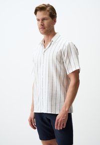 Korte mouw, knoopsluiting shirt in lichtbeige linnen met verticale marineblauwe strepen, voorzien van een kraag en een ontspannen pasvorm. Draagt bij donkere shorts.