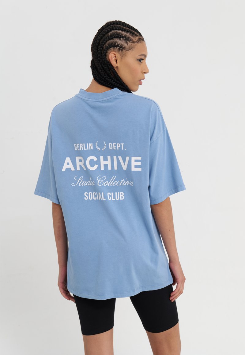 Lys blå, overdimensjonert t-skjorte med hvit tekst på baksiden: "BERLIN DEPT. ARCHIVE Studio Collection SOCIAL CLUB." Laget av myk stoff.
