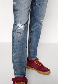 Slitna blå denimjeans med hål och färgstänk, parat med röda sneakers som har gula skosnören och en beige sula.