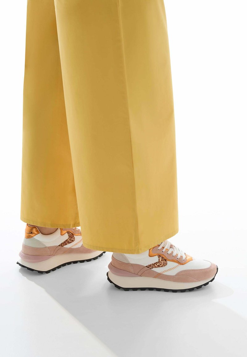 Baskets blanches et beiges avec des accents orange et des détails en paillettes, associées à un pantalon large jaune. Matériaux texturés et silhouette distincte.
