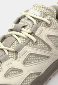 Helle beigefarbene Sneaker mit einem Obermaterial aus Mesh, glatten Wildlederakzenten, cremefarbenen Schnürsenkeln und auffälligen geometrischen Ausschnitten an den Seiten; strukturierte Oberfläche.