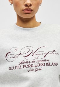 Pelēka džemperis ar ribotu apkakli. Uz tā ir izšūti bordo krāsas vārdi: "East Hampton," "Atelier de creation," un "South Fork, Long Island."